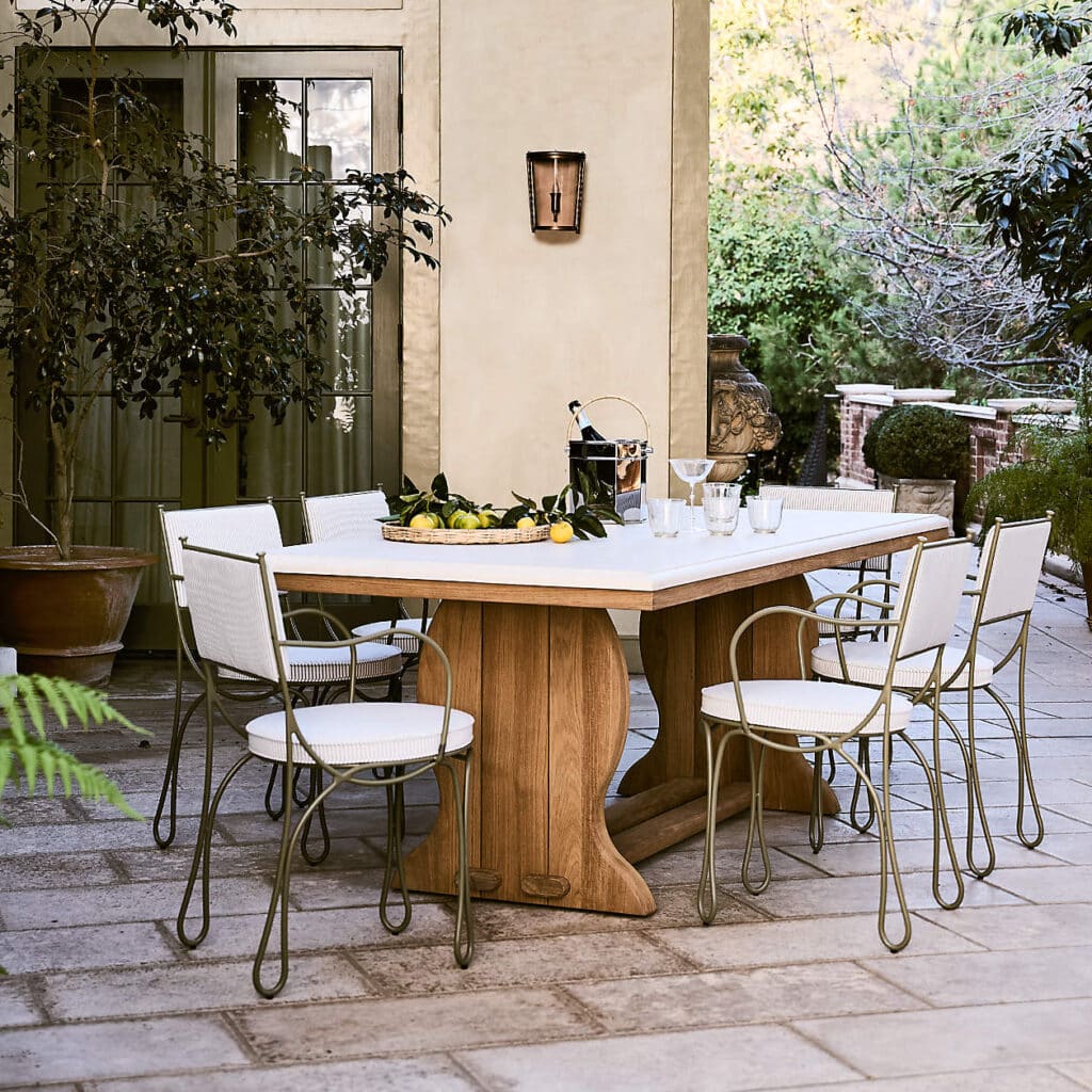 Elegant outdoor dining setup with wooden table, white chairs, and a bowl of lemons on a patio surrounded by greenery.