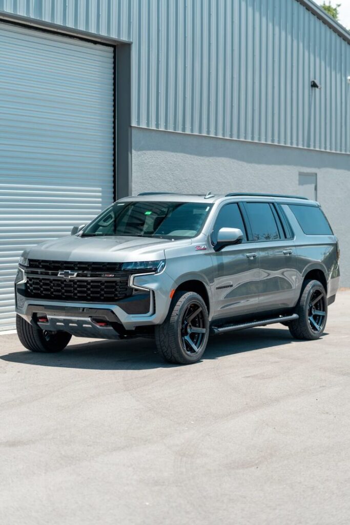 CHEVROLET SUBURBAN 4PLAY WHEELS 4P55 RIMS 24X12 33X12_5X24 RBP TIRES STOCK SUSPENSION iPHONE PICS - 4PLAY Wheels Silver SUV parked in front of a metal garage door under a clear sky. | Sky Rye Design Silver SUV parked in front of a metal garage door under a clear sky.