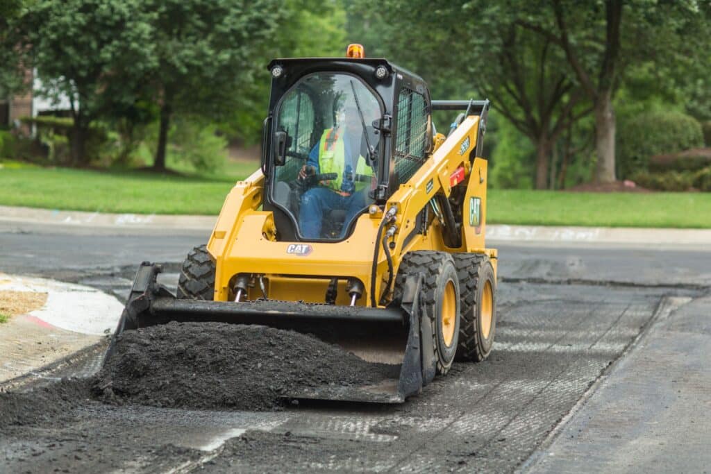Yellow skid steer loader paving an asphalt road on a sunny day, surrounded by green trees and grass.