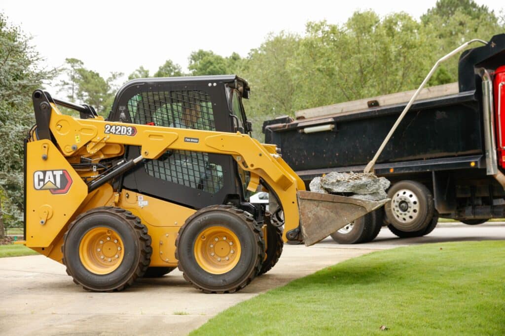 Yellow skid steer loader transporting rocks next to a dump truck on a grassy construction site.