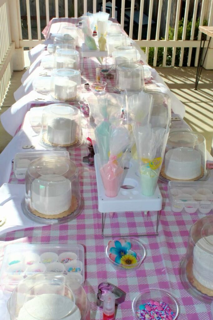 Cake decorating setup with white cakes, icing, and sprinkles on a pink checkered tablecloth outdoors.