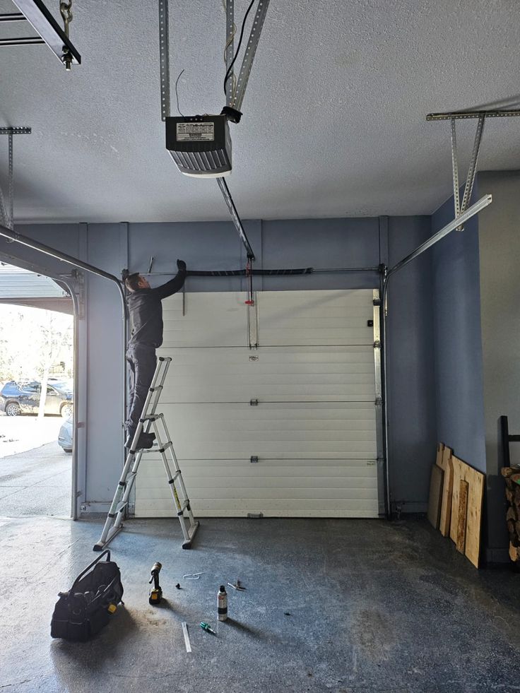 Technician on a ladder repairing a garage door opener inside a garage. Tools and equipment are visible on the floor.