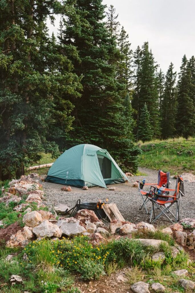 Green tent in a forest campsite with chairs and a fire pit nearby, surrounded by trees and rocks.
