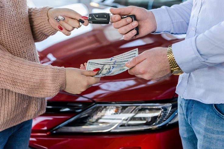 Exchange of car keys and cash in front of a red vehicle, symbolizing car sale or purchase transaction.