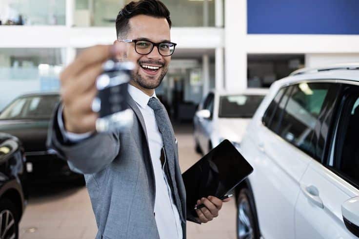 Smiling car salesman holding keys and a tablet, standing next to a white vehicle in a dealership showroom.