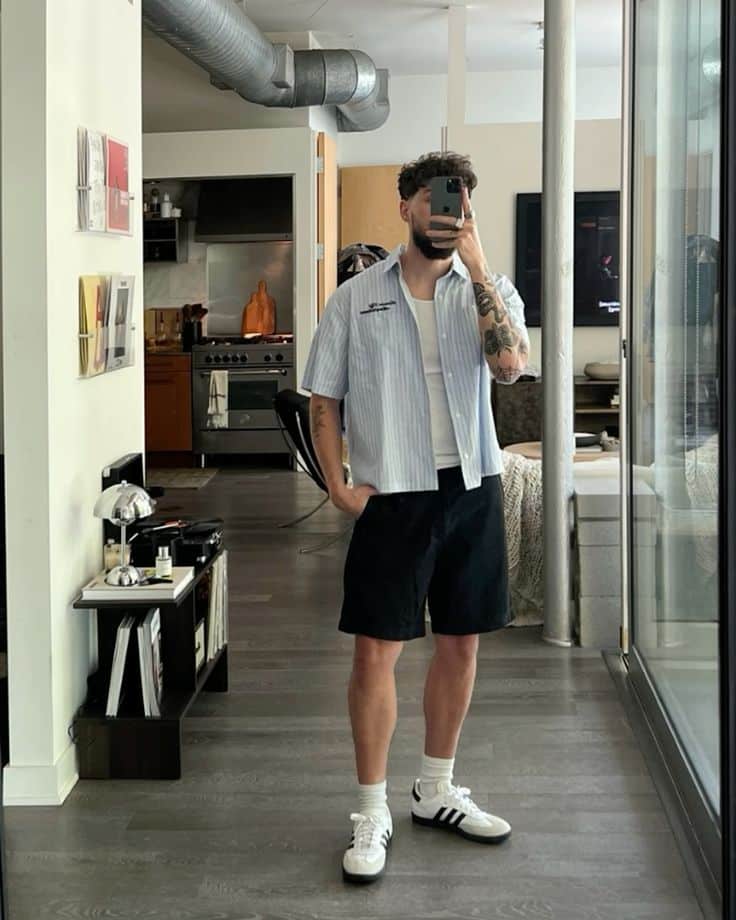 Man taking mirror selfie in casual outfit with striped shirt, black shorts, and sneakers in modern apartment.