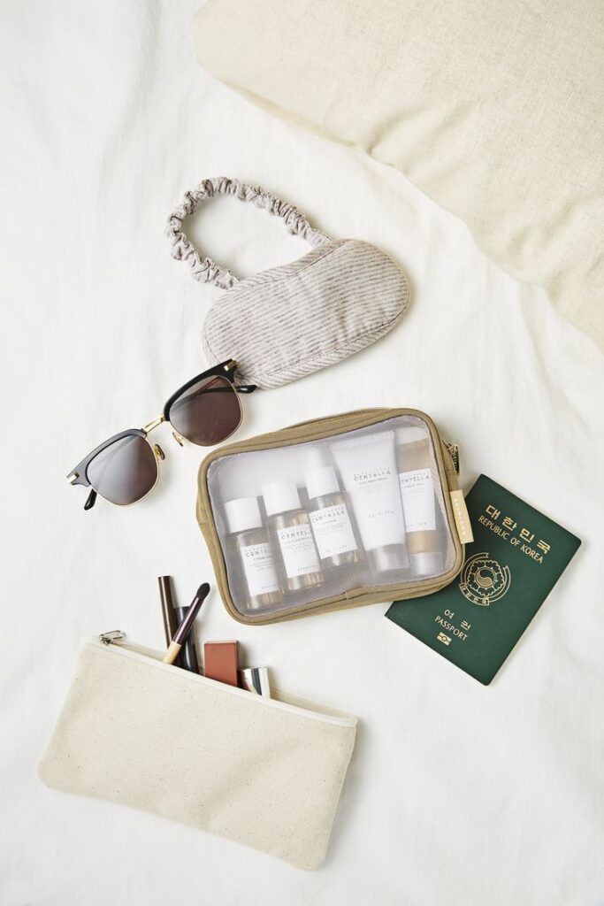 Travel essentials on bed: eye mask, sunglasses, skincare pouch, makeup bag, Korean passport. Ideal for a stylish getaway.