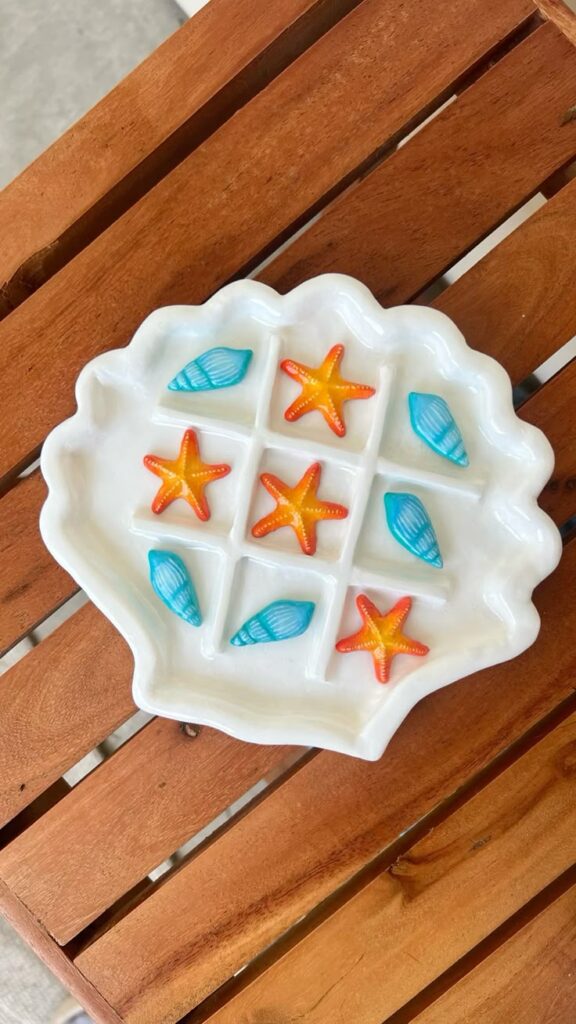 Shell-shaped tic-tac-toe board with colorful starfish and seashells on a wooden table.