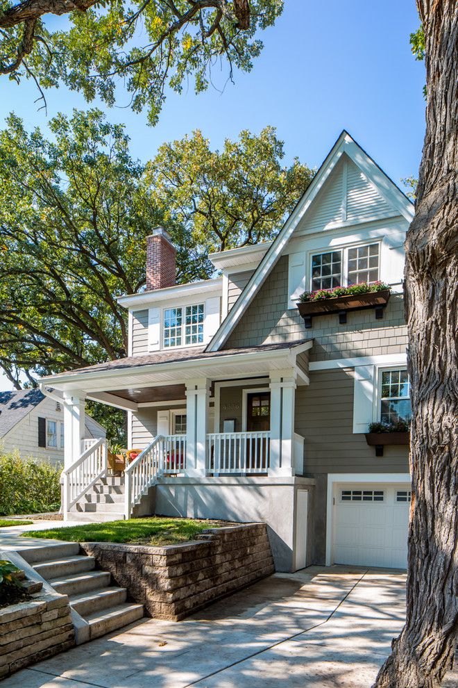Charming suburban house with porch, chimney, and garage, surrounded by trees on a sunny day.