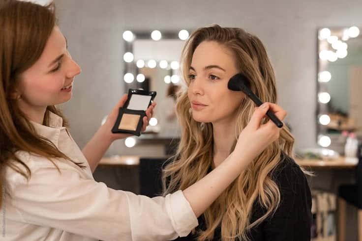 Makeup artist applying blush to a woman's face in a well-lit studio. Makeup