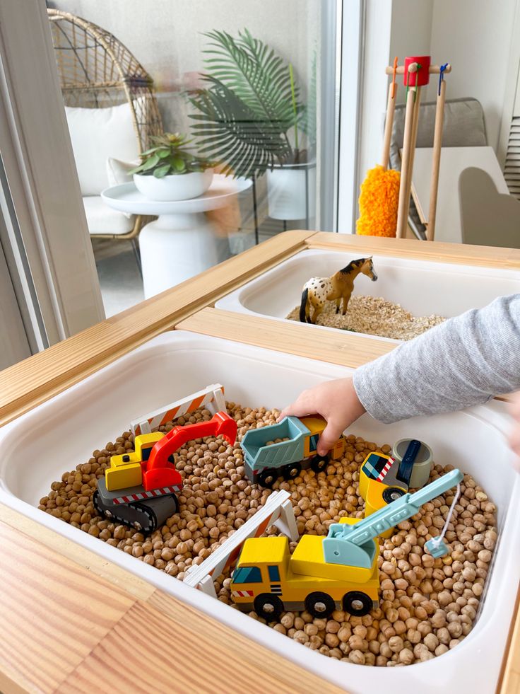 Child playing with toy construction vehicles in a sensory bin filled with chickpeas indoors.
