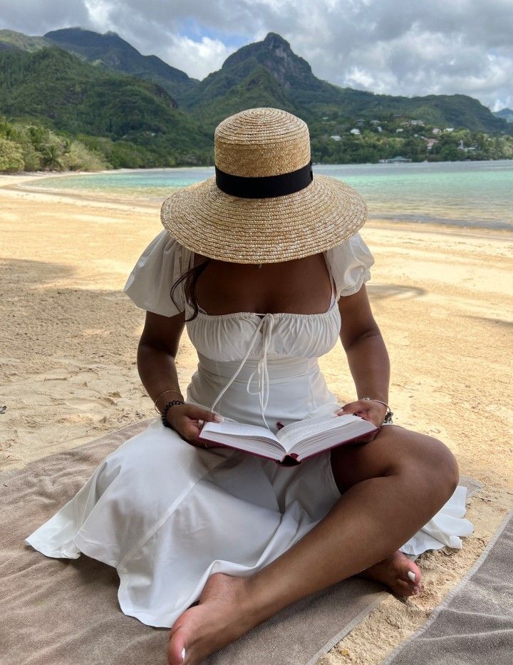 Woman in straw hat reading a book on tropical beach with mountains in the background.
