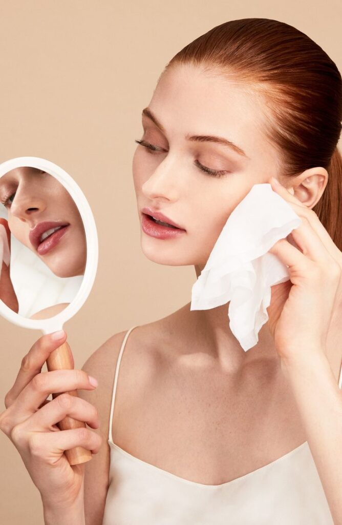 Woman using facial wipe while holding mirror, showcasing skincare routine on neutral background.