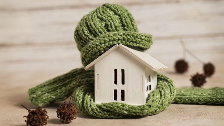 Miniature house wrapped in a warm green scarf, symbolizing home insulation and energy efficiency in winter.