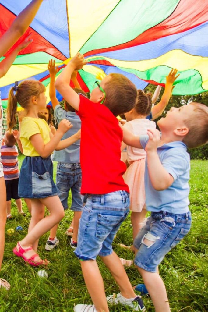 Children playing with a colorful parachute outdoors, enjoying group fun and games in a park setting.
