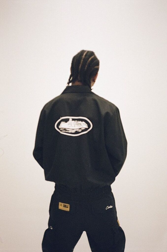 Man with braided hair wearing a black jacket featuring a logo on the back, facing away, on a plain background.