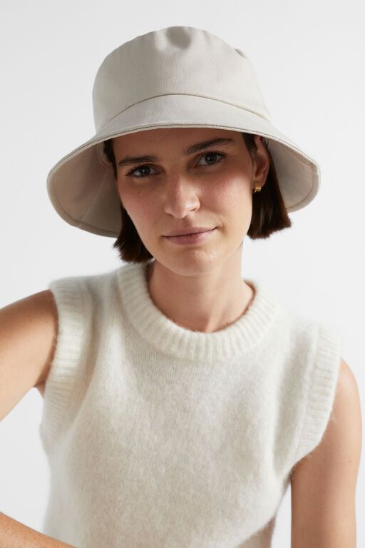 Person wearing a beige bucket hat and sleeveless sweater, smiling softly against a white background.