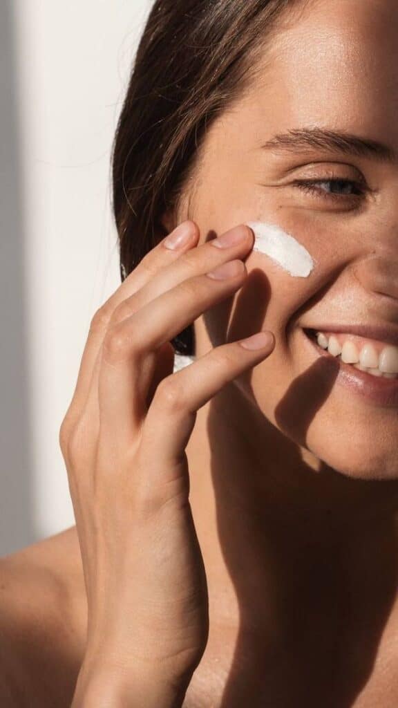 Woman applying sunscreen to her face, smiling in natural sunlight. Skincare routine for healthy, glowing skin.