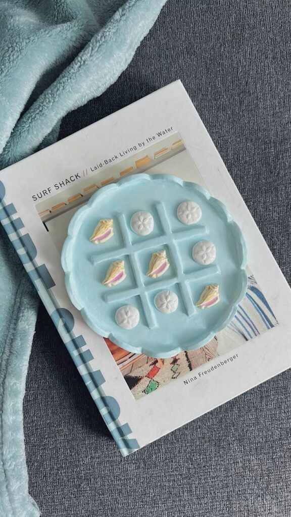 Tic-tac-toe game on a blue board with seashells and pastries atop a Surf Shack book on a cozy blanket.