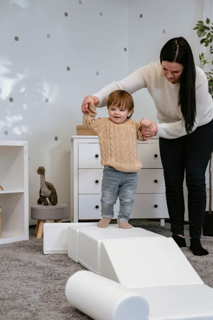 Toddler in beige sweater practices balance on a foam ramp, assisted by adult in cozy room.
