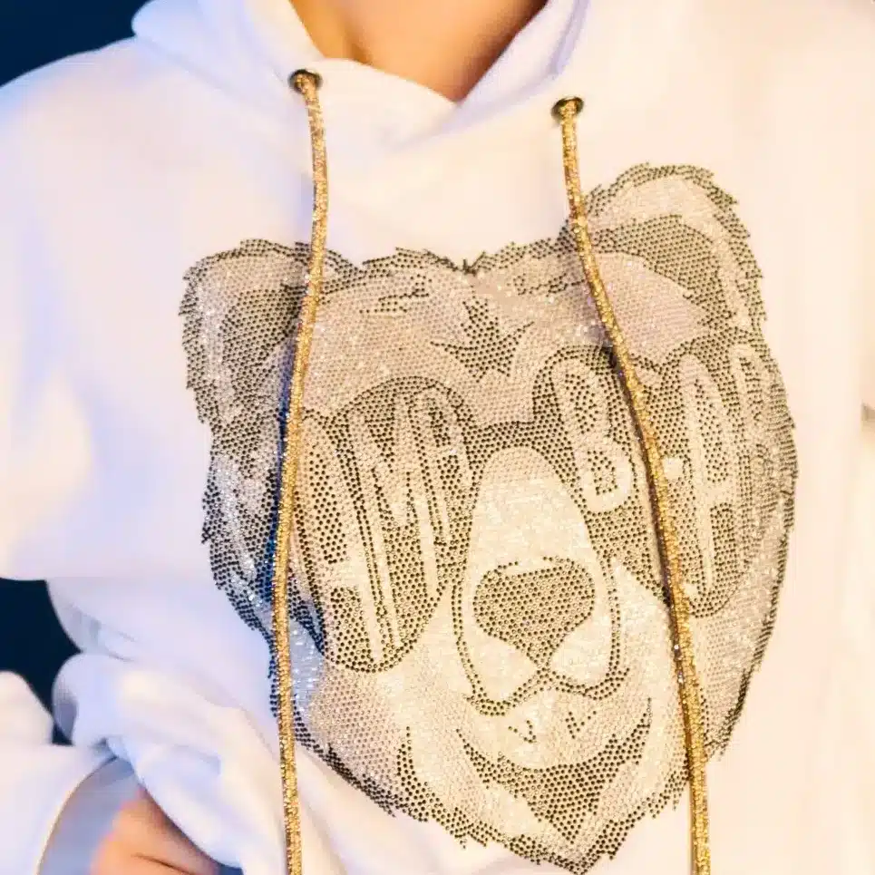 Woman in white hoodie featuring a rhinestone bear design and gold drawstrings, posing against a dark background.