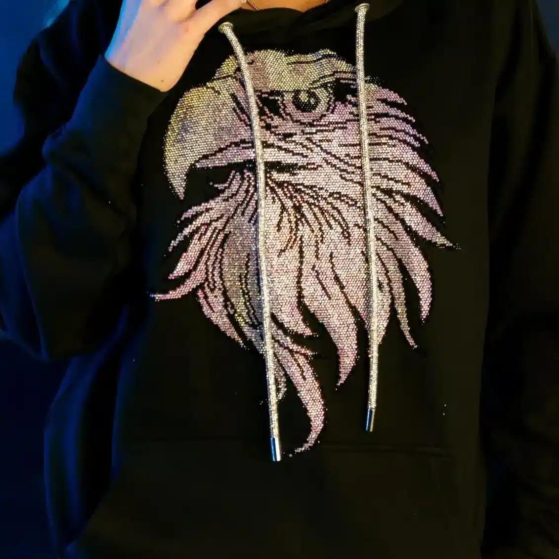 Person wearing a black hoodie with a sparkling eagle design, standing against a dark blue background.