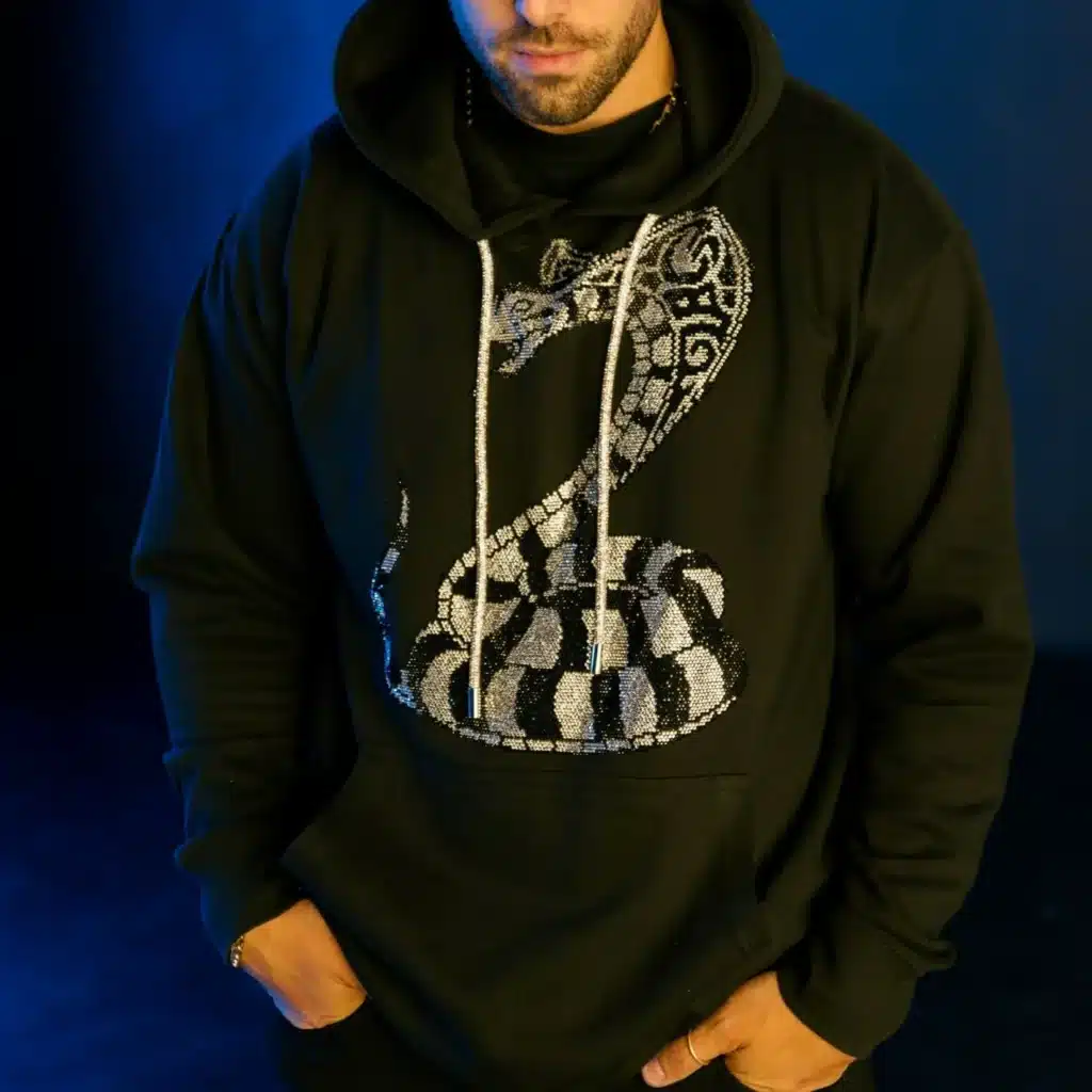Man wearing a black hoodie with a stylized cobra design, standing against a dark blue background.