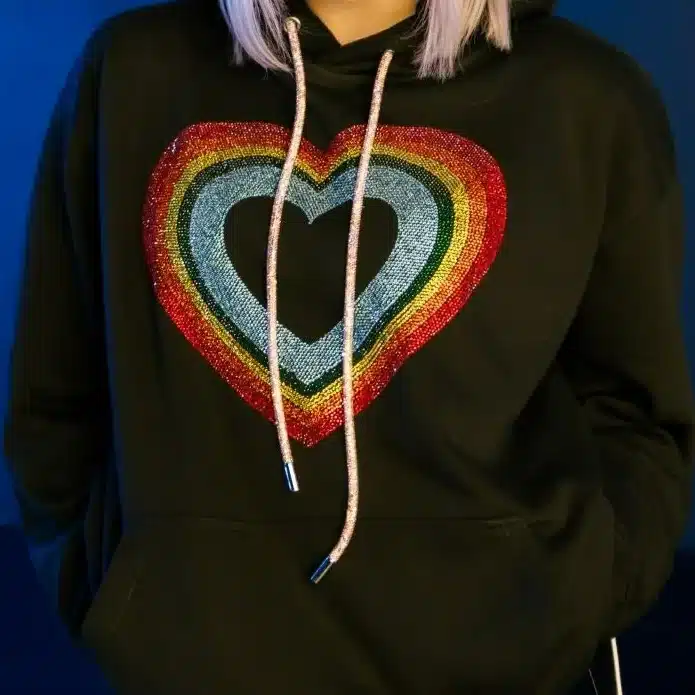 Person wearing a black hoodie with rainbow heart design, smiling against a blue background.
