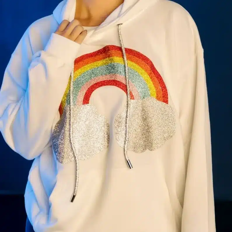 DSC_7881 Person wearing a white hoodie with rainbow and cloud design against a blue background. | Sky Rye Design Person wearing a white hoodie with rainbow and cloud design against a blue background.