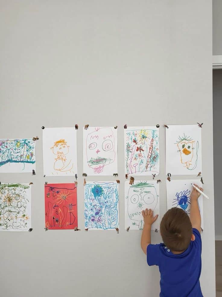 Child drawing on wall, adding artwork to a display of colorful children's drawings, creativity in progress.