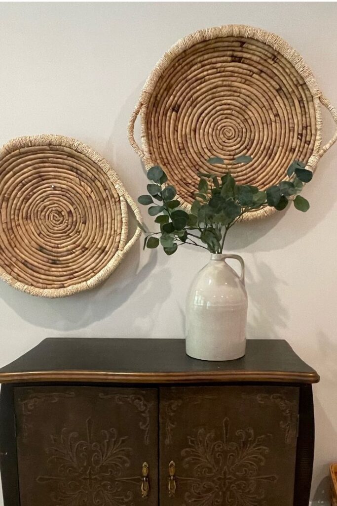 Woven wall baskets above a vase with green foliage, placed on ornately carved dark wooden cabinet.