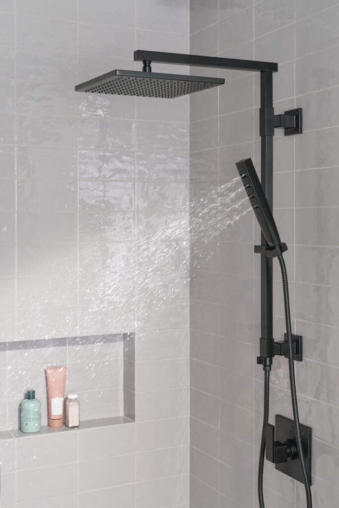 Modern black showerhead spraying water in a tiled bathroom with bath products on shelf.