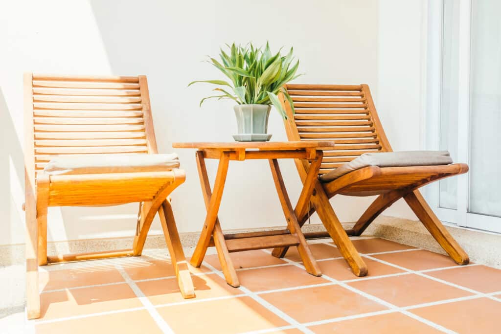 Two wooden chairs and a table with a potted plant on a sunlit patio with tile flooring.