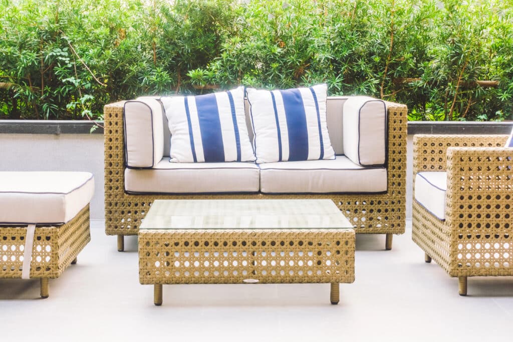 Depositphotos_105692064_XL Outdoor wicker patio furniture with striped cushions and glass table, set against lush greenery. | Sky Rye Design Outdoor wicker patio furniture with striped cushions and glass table, set against lush greenery.