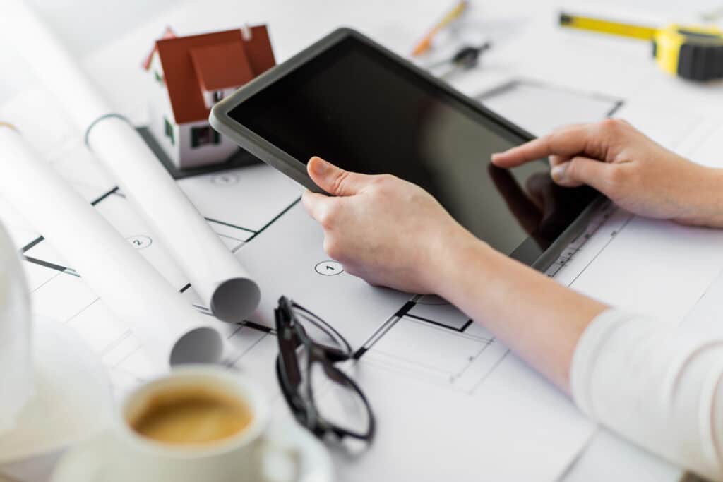 Architect reviewing house plans on tablet with model, blueprints, coffee, and glasses on desk.