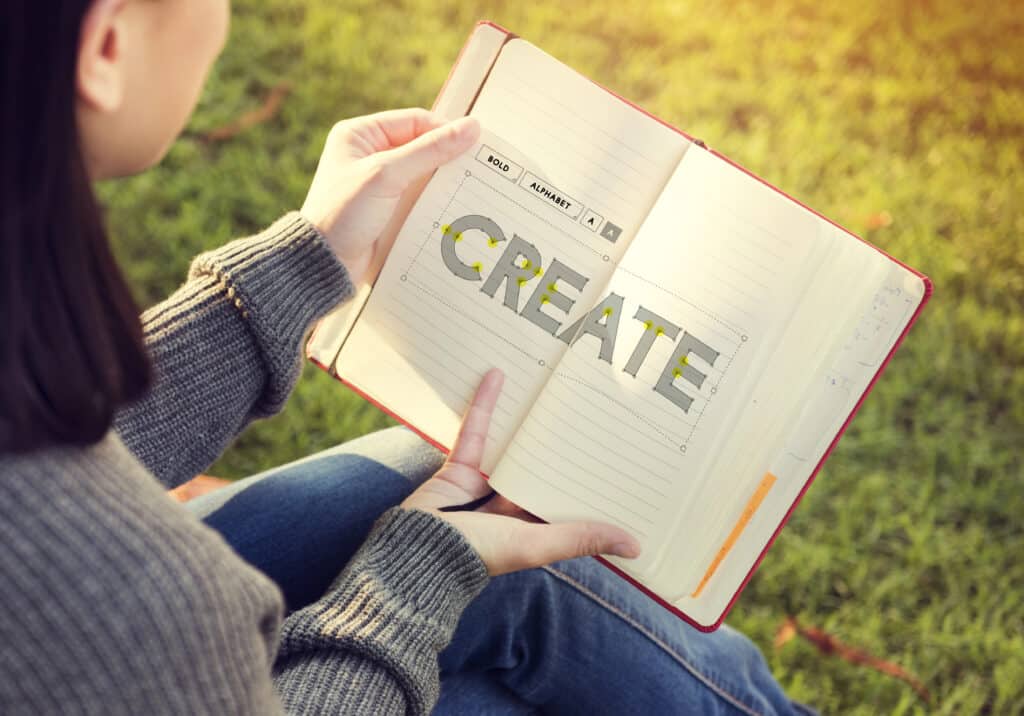 Person holding notebook with the word 'Create' on a grassy background, emphasizing creativity outdoors.