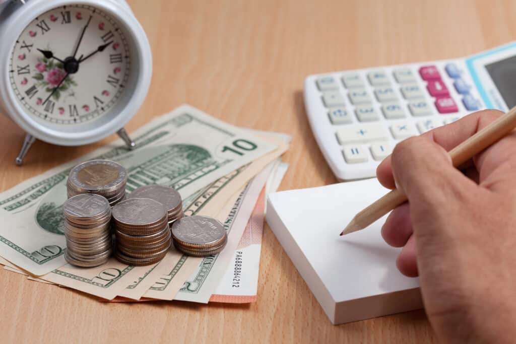 Coins and bills next to a clock, calculator, and hand writing on pad, symbolizing budgeting and time management.