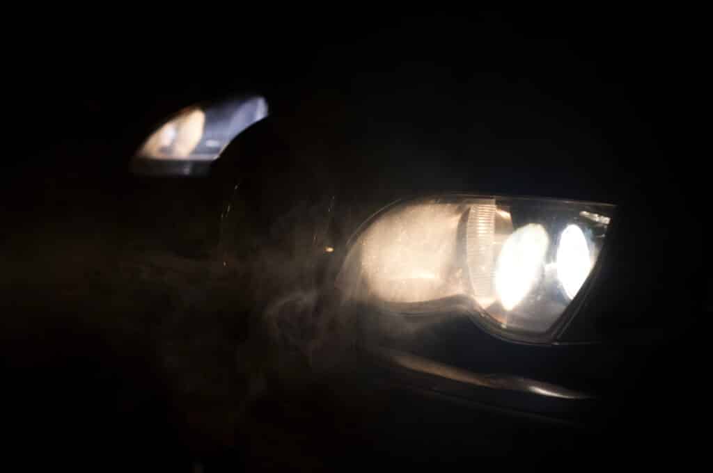 Close-up of a car's illuminated headlights in darkness, with visible smoke and a moody atmosphere.