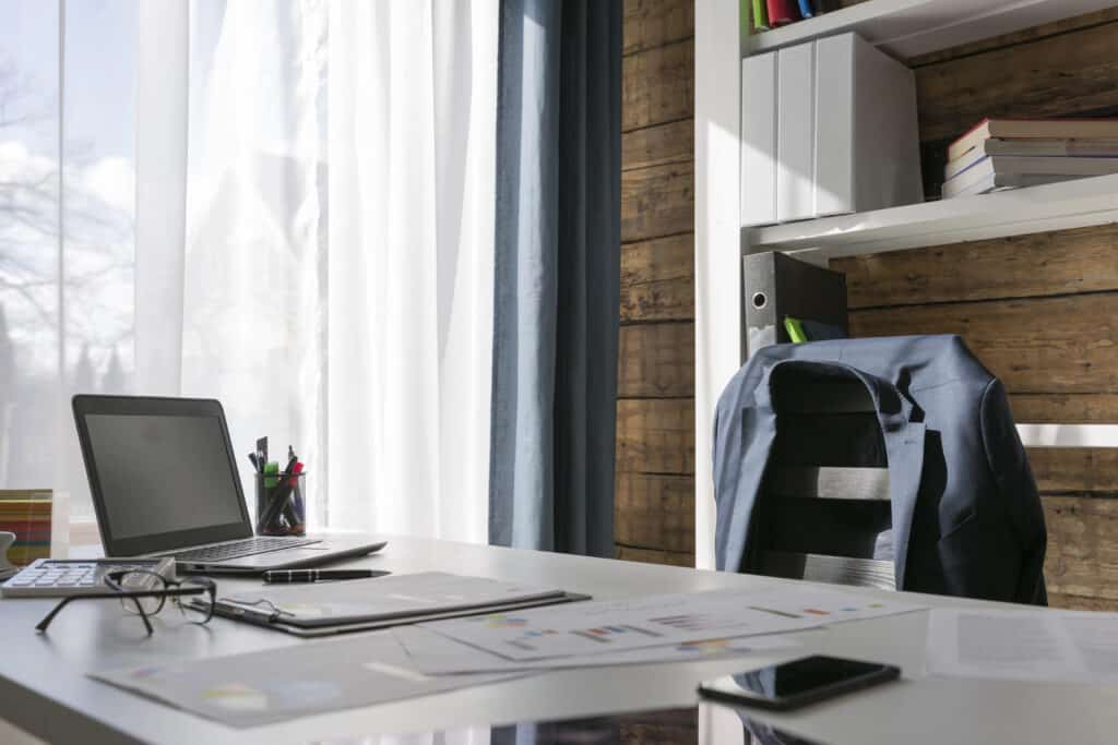 Modern home office with laptop, documents, glasses, and a jacket on the chair, bathed in natural light.