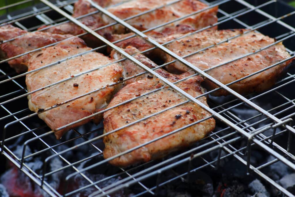 Grilled pork chops seasoned and cooking over hot charcoal on a metal grill rack.