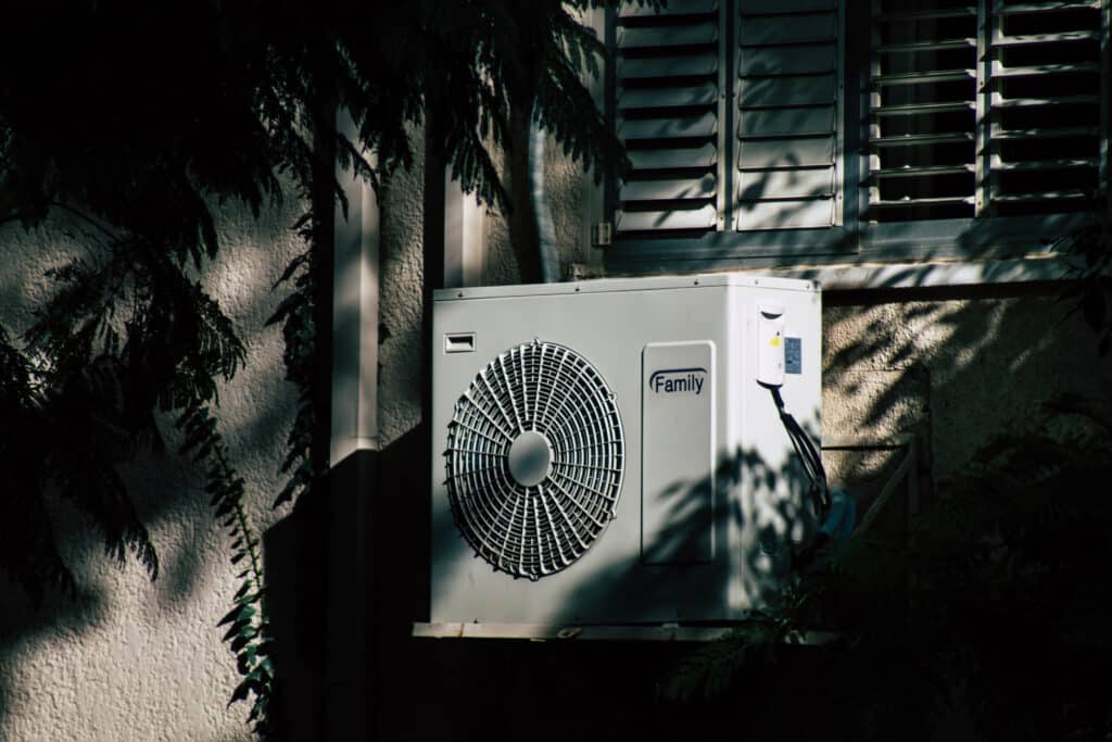 Outdoor air conditioning unit mounted on a house wall, surrounded by foliage shadows.