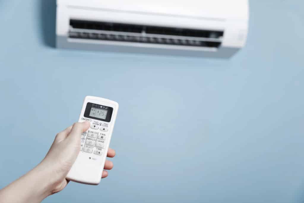 Hand holding remote control to adjust air conditioner temperature in a room, with a blue wall background.