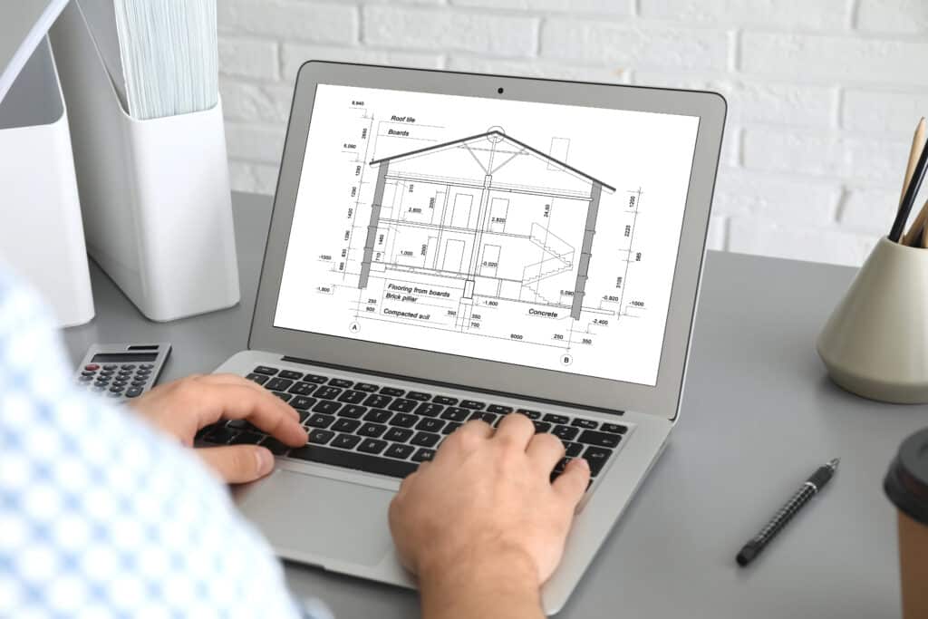 Depositphotos_377454510_XL Person reviewing architectural house plans on a laptop in an office setting. | Sky Rye Design Person reviewing architectural house plans on a laptop in an office setting.