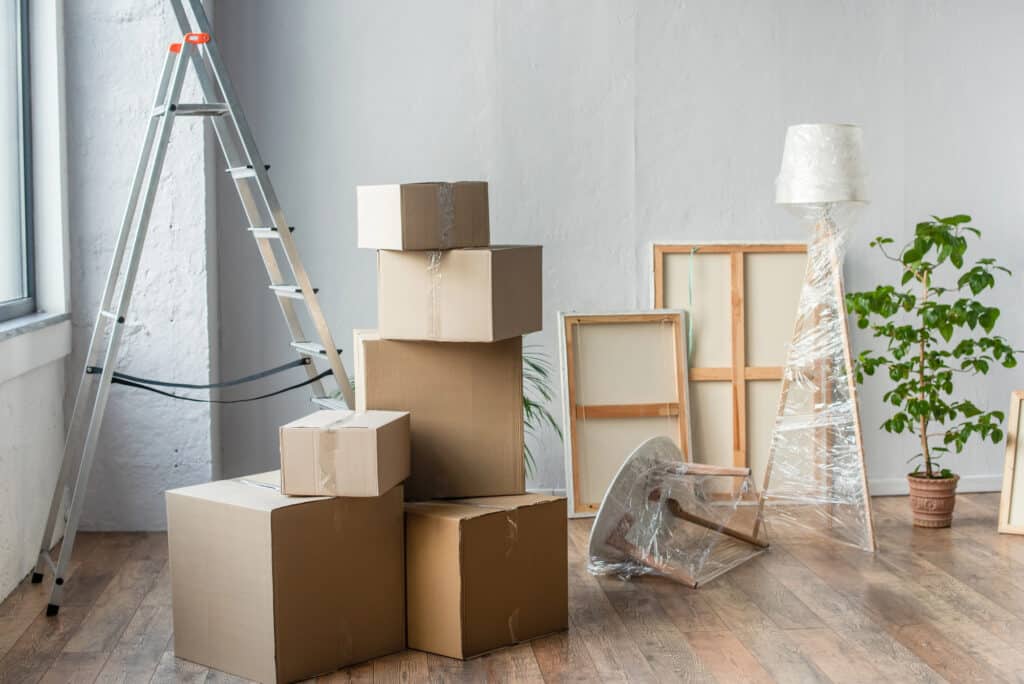 Moving day scene with stacked cardboard boxes, ladder, and wrapped furniture in a bright, empty room.