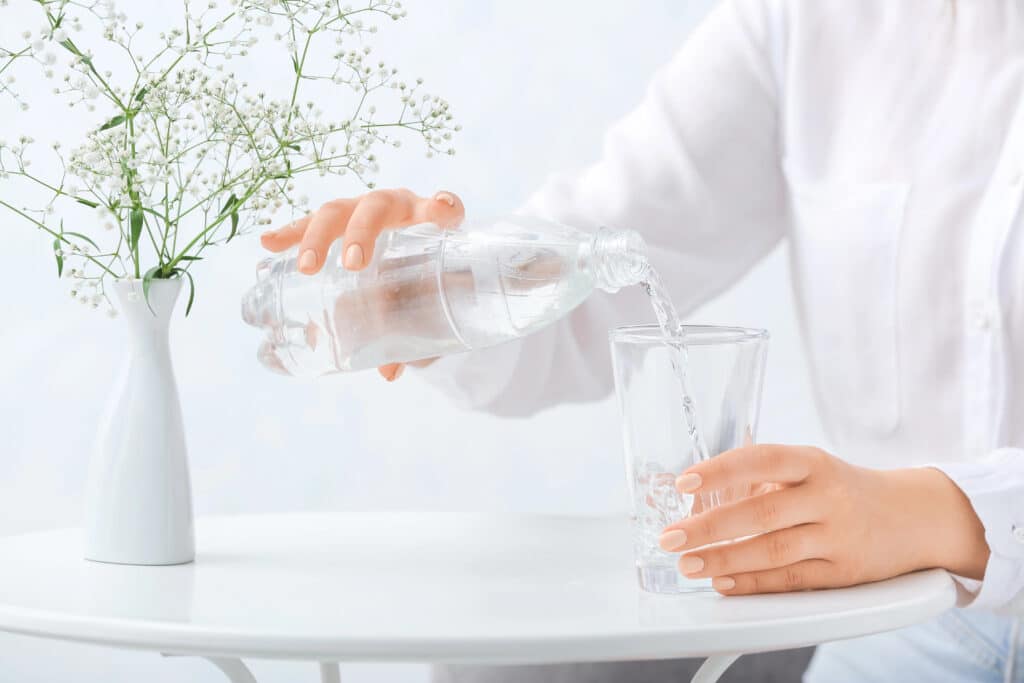 Depositphotos_432302360_XL Pouring water into a glass on a white table beside a vase of small white flowers, promoting hydration and simplicity. | Sky Rye Design Pouring water into a glass on a white table beside a vase of small white flowers, promoting hydration and simplicity.