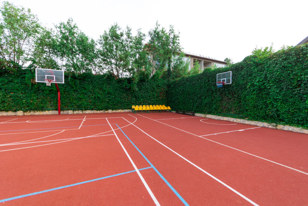 Outdoor basketball court with red surface, green hedge, and yellow seating, surrounded by tall trees.