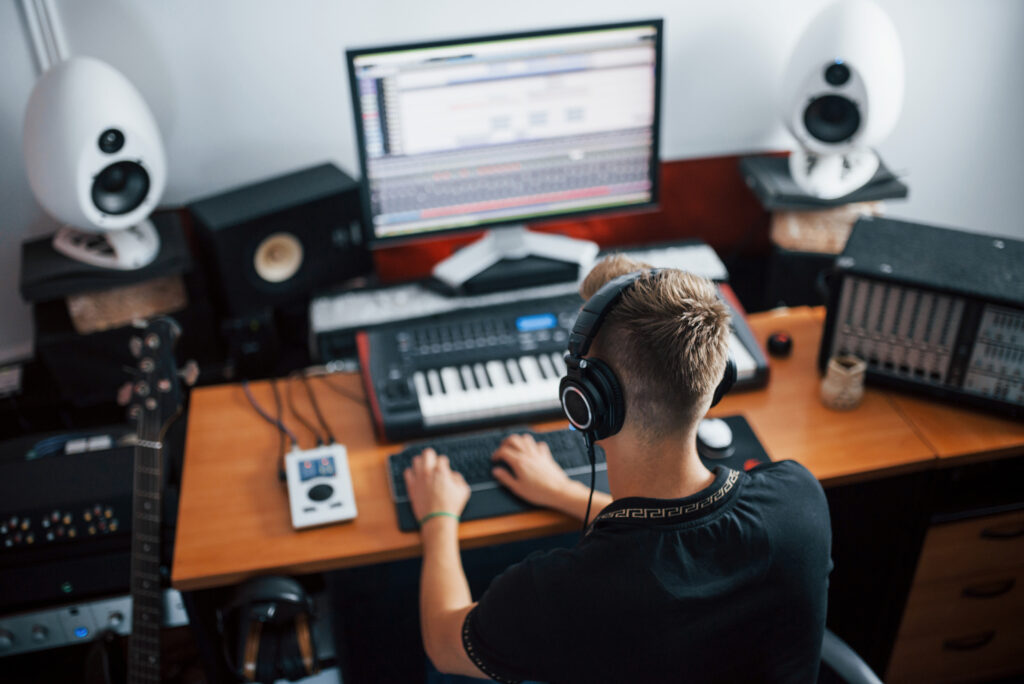 Sound engineer in headphones working and mixing music indoors in the studio Person at music production desk with computer, keyboard, headphones, and speakers in a studio setup. | Sky Rye Design Person at music production desk with computer, keyboard, headphones, and speakers in a studio setup.