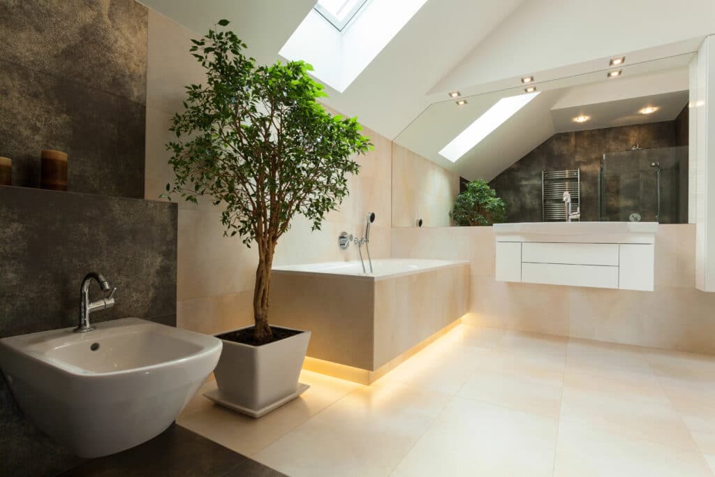 Modern bathroom with a skylight, white fixtures, large bathtub, potted plants, and elegant lighting.