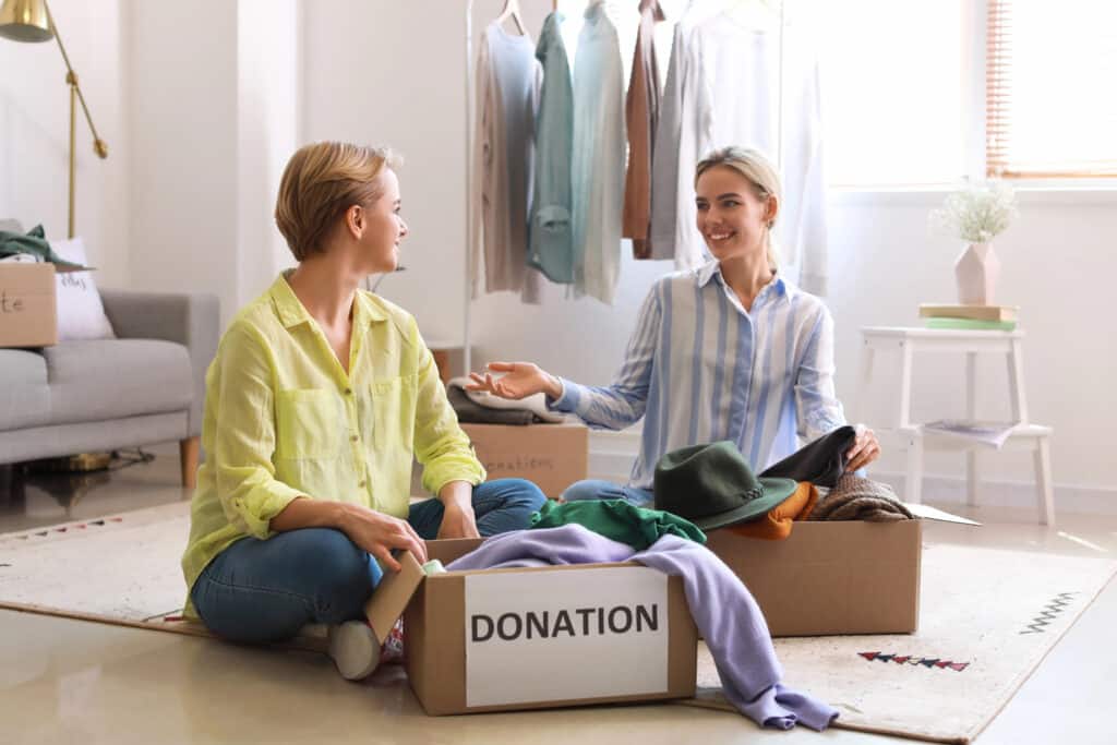 Two women sorting clothes for donation in a bright room, with a labeled box and garments on racks.
