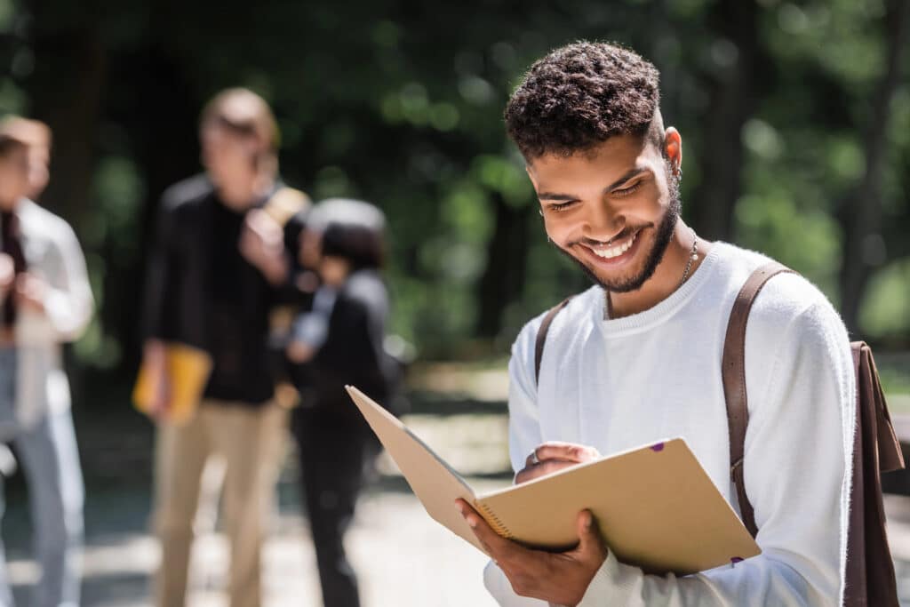 Man smiling while reading notebook outdoors, with blurred group of people in the background.